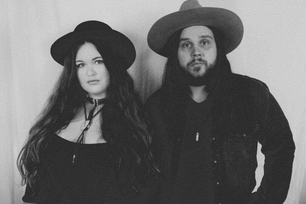 Man and a woman stand looking at the camera wearing a hat.