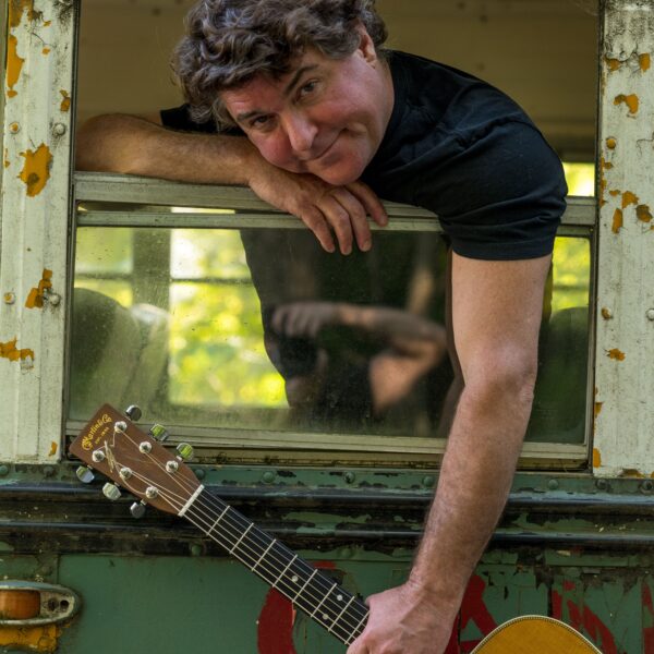 Keller Williams, brunette man, hanging his arm out of a bus window with a guitar in his hand.