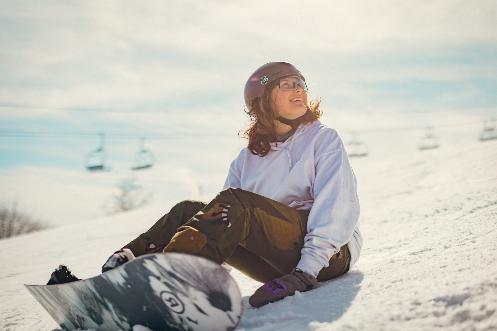 Girl strapped into a Burton snowboard looking up the ski slopes smiling, sitting down strapped in.